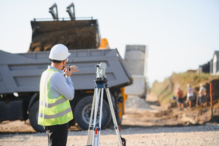 Engineer working at a construction site, using surveying equipment and a walkie talkie while a dump...