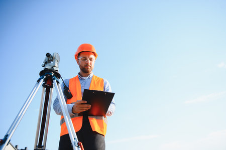 Professional surveyor in safety vest and hard hat using a theodolite for land measurement against a...