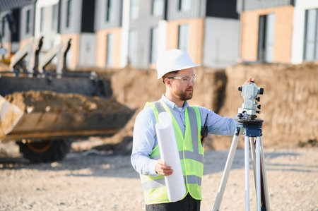 Male surveyor wearing hard hat and safety vest, using a theodolite for land measurement and planning...