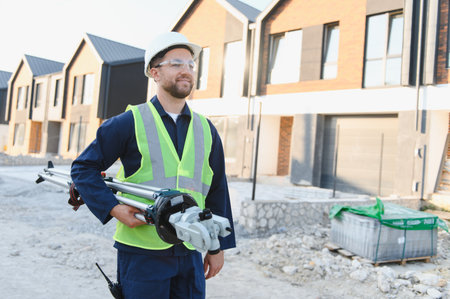 Male surveyor wearing hard hat and safety vest, holding surveying equipment, with new residential...