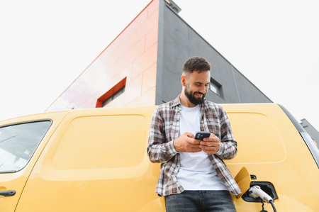 Delivery driver checking smartphone while charging electric van outside a sleek, modern building,...