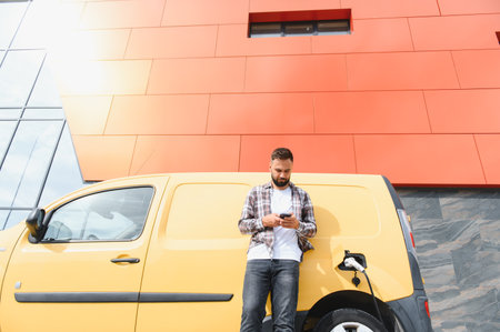 Delivery driver using smartphone while his electric van is charging in front of modern building