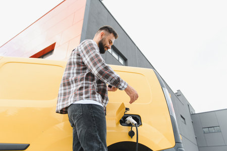 Delivery driver engaging with a mobile phone app while charging an electric van at a charging...