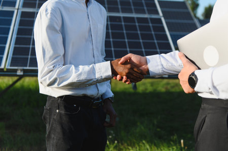 Two diverse businessmen shaking hands against solar panels, symbolizing partnership and agreement in...