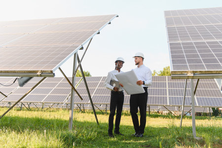 Two diverse engineers discussing blueprints and planning green energy solutions at a large solar...