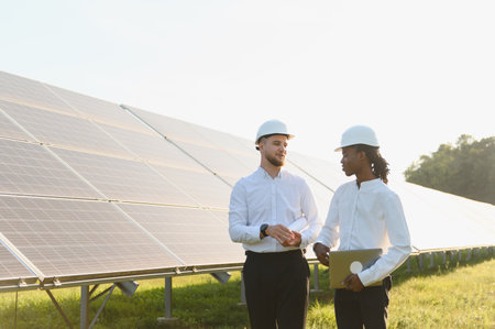 Two diverse engineers in hard hats and white shirts discussing plans and holding a laptop near solar...