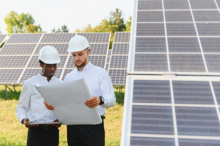 Diverse engineers in hard hats reviewing a blueprint and tablet at a solar energy farm, discussing...
