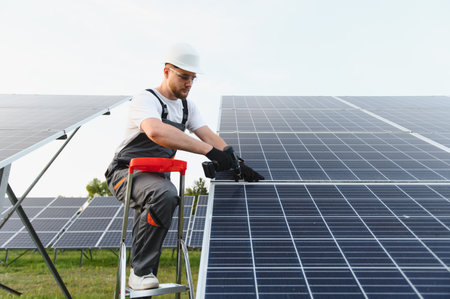 Man in overalls and safety helmet working on solar farm, installing photovoltaic panels for green...