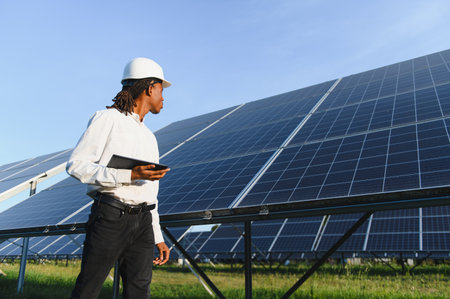 African american engineer wearing hard hat, holding tablet, inspecting rows of solar panels at a...