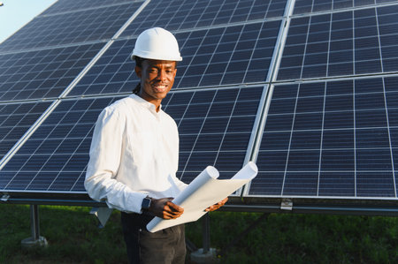 African american engineer wearing a hard hat, smiling and holding blueprints in front of solar panels. Renewable energy conceptの写真素材
