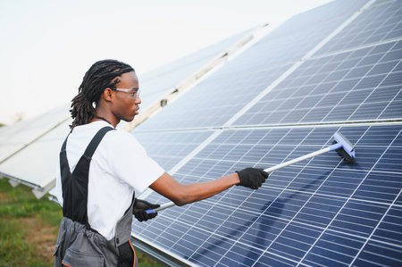 Male worker with safety goggles cleaning a large solar panel array, ensuring optimal performance for renewable energy generationの写真素材