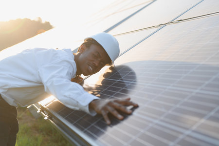 African american man in hard hat examining photovoltaic panel during sunset, ensuring renewable energy system performanceの写真素材