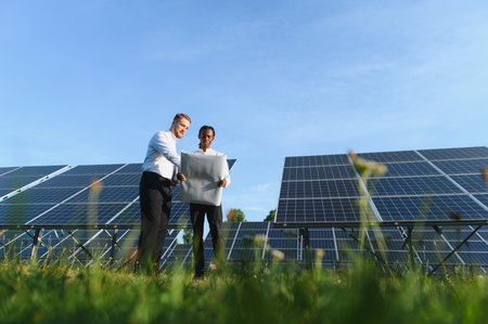Two engineers reviewing a blueprint plan while standing among rows of solar panels, collaborating on...