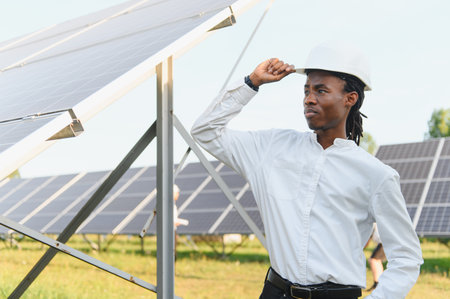 Young black man in hard hat inspecting solar panels, representing sustainable technology and clean...