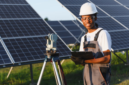 Black engineer wearing a hard hat and safety glasses, surveying a solar panel farm using a theodolite and clipboardの写真素材