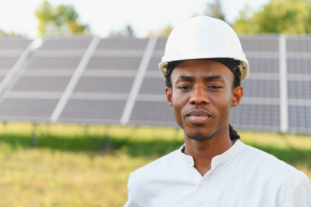 Young black man wearing a hard hat, standing in front of solar panels. Representing green energy and sustainable technologyの写真素材