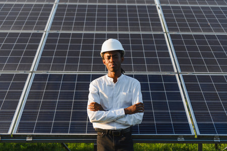 Young man wearing a hard hat, arms crossed, standing proudly at a solar power plant, representing renewable energyの写真素材