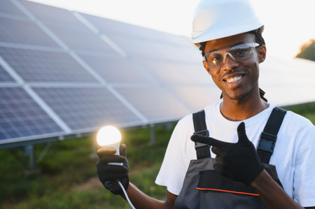 African american engineer holding a glowing light bulb, confidently pointing, with solar panels in the background, representing clean energyの写真素材
