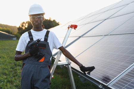 African american technician holding a power drill, installing solar panels on a green field. Concept of green energy and sustainable futureの写真素材