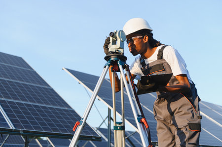 African American engineer operating surveying equipment, precisely measuring and documenting solar panel installation at a renewable energy facilityの写真素材