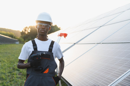 African american technician wearing hard hat, safety glasses, and overalls, standing proudly beside solar panels at sunsetの写真素材