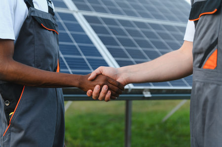 Technicians from different backgrounds shaking hands in front of solar panels, symbolizing...