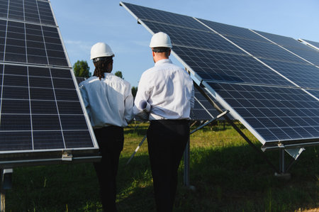Two diverse engineers in hard hats examining solar panels, planning and managing a sustainable green...
