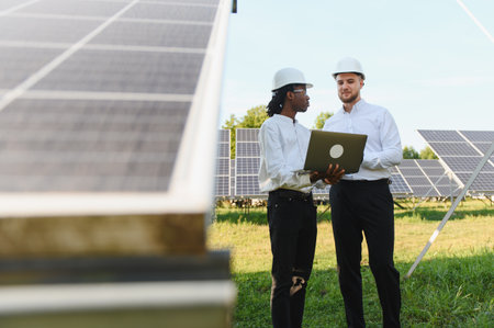 Two engineers using a laptop, discussing a power plant project with many solar panels collecting...