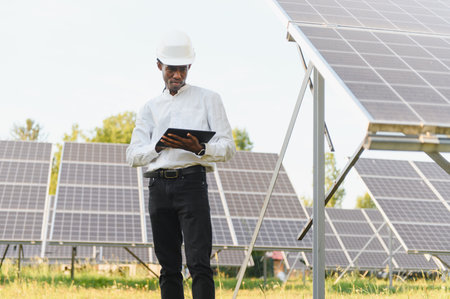 Young black man engineer wearing hard hat standing in a solar panel field, holding digital tablet...