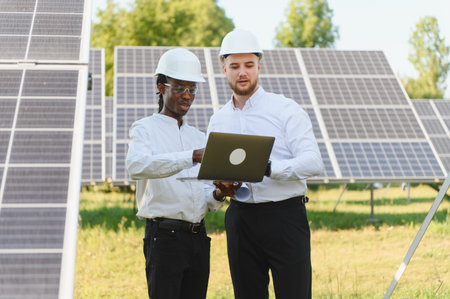 Two diverse engineers wearing hard hats reviewing plans on a laptop at a solar power plant,...