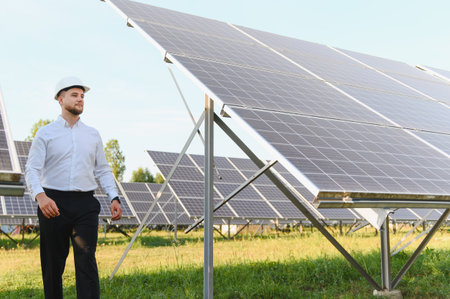 Young engineer in hard hat inspecting solar panel field, ensuring efficient operation and...