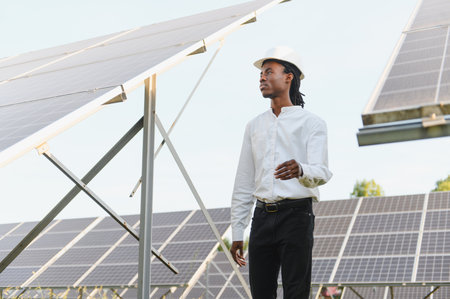Young black engineer in hard hat inspecting solar panels at a daytime renewable energy power plant, sustainable tech siteの写真素材