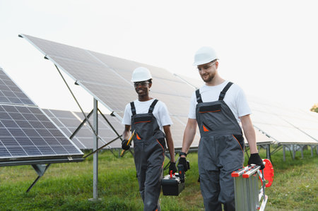 Diverse engineers carrying tools and ladder, walking past rows of solar panels at a renewable energy...