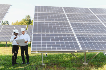 Two engineers are reviewing schematics at a solar energy farm, collaborating on renewable clean...