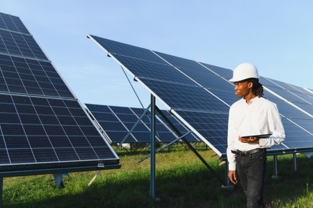 Engineer wearing a hard hat and holding a tablet, standing in a large solar panel field representing...