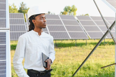 African american engineer wearing a hard hat, holding a digital tablet, standing near solar panel...