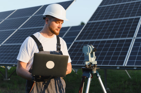 Engineer wearing hardhat and overalls checking data on a laptop, standing next to a total station...
