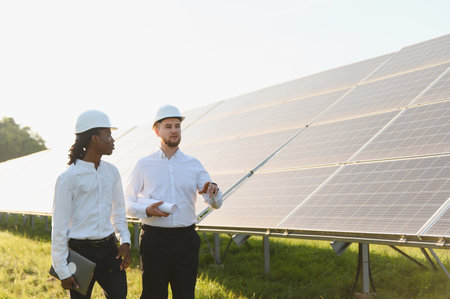 Two engineers, one black, one caucasian, inspecting a large solar panel installation, discussing...