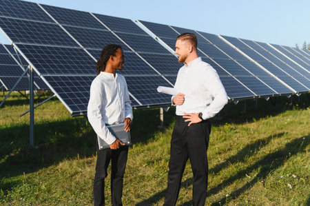 Two diverse engineers standing in front of solar panel array, discussing sustainable energy concepts...