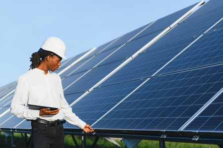 African american man wearing hard hat and holding a tablet, checking large solar panels for...