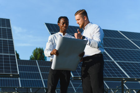 Two diverse engineers collaborating, reviewing a blueprint at a solar power plant, symbolizing clean...