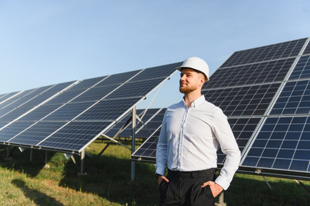 Man in hard hat and shirt standing proudly in front of a large solar panel array, representing clean...