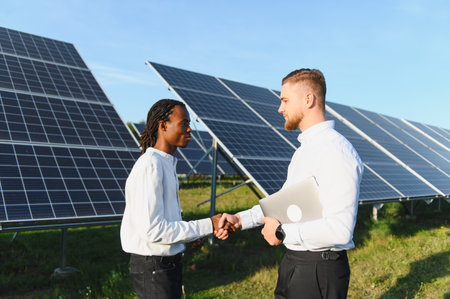 Two diverse businessmen shaking hands near solar panels, symbolizing partnership, renewable energy,...