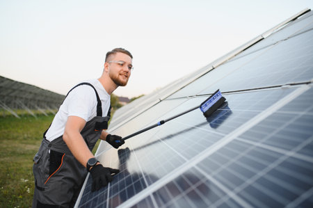 Young man in overall and safety glasses cleaning a solar panel field for efficiency and sustainable energy productionの写真素材