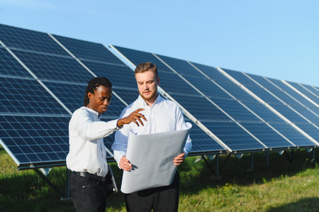 Two diverse engineers discussing a blueprint, planning sustainable energy solutions at a solar panel...