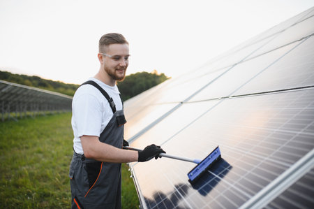 Young man in overalls and goggles cleaning solar panels with a squeegee, performing maintenance at a...