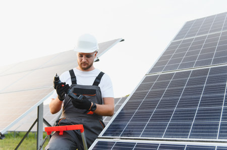 Young man wearing a hardhat and protective glasses, preparing a drill while working on a solar power...