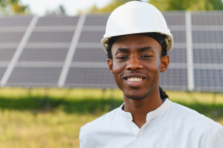 Smiling african american engineer wearing a hard hat, standing in front of solar panels. Concept of clean energy and sustainabilityの写真素材