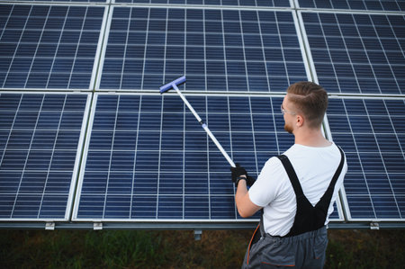 Male worker performing maintenance, cleaning solar panels with a squeegee tool to ensure optimal...