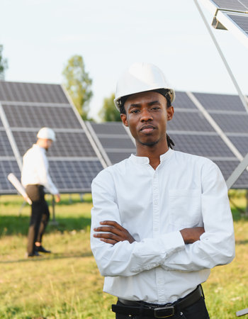 African american engineer in a hard hat standing with arms crossed, another worker inspecting solar panels in backgroundの写真素材